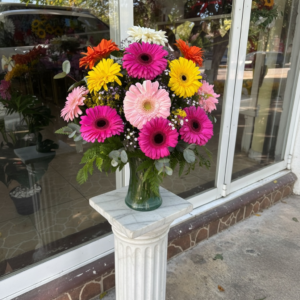 Colorful bouquet of gerbera daisies in pink, yellow, orange, and white, arranged in a green vase on a marble pedestal outside a storefront window.