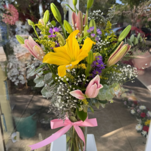 Bouquet of yellow lilies, pink lilies, purple blooms, and baby's breath in a glass vase with a pink ribbon.