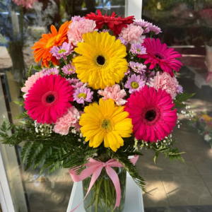 Colorful bouquet of gerbera daisies in a glass vase with a pink ribbon on a white pedestal by a glass door.
